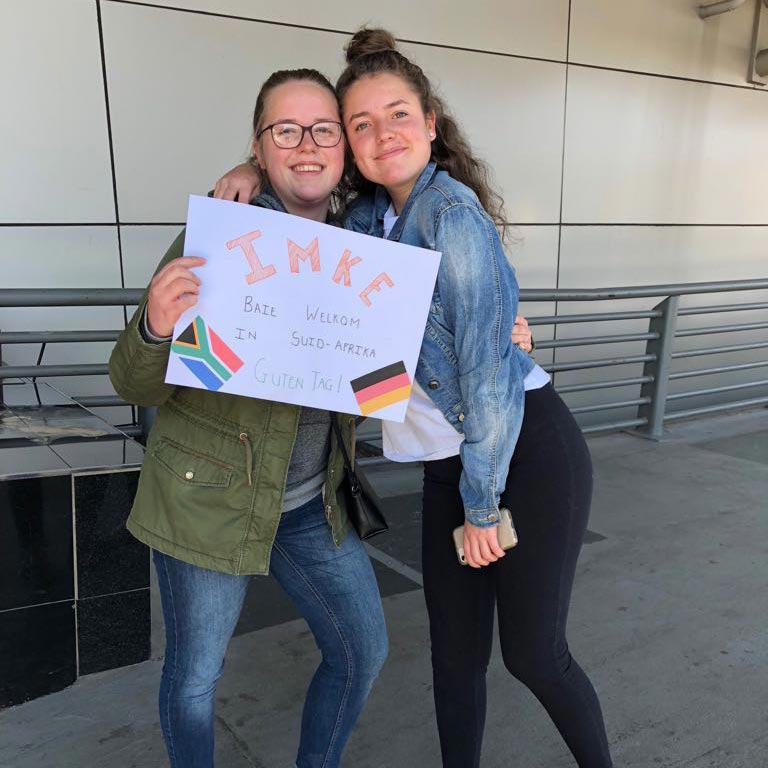 Zwei Freundinnen halten Arm in Arm ein selbst gebasteltes Namensschild hoch