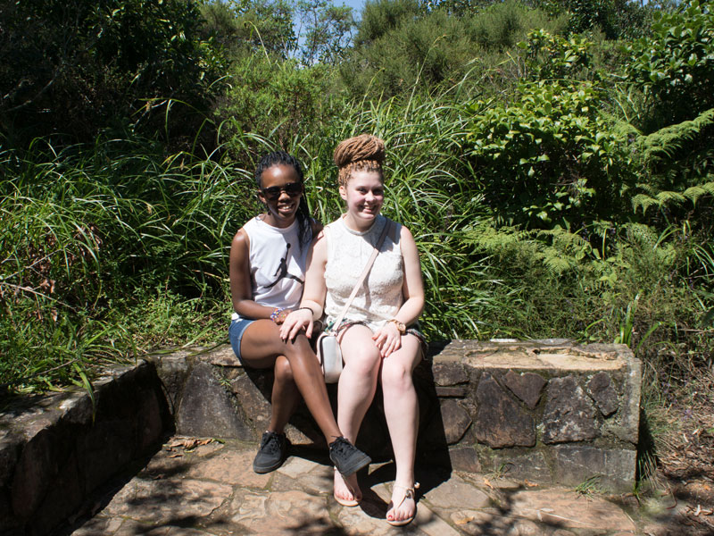 Ein deutsches und ein südafrikanisches Mädchen sitzen zusammen freudig auf einer kleinen Mauer