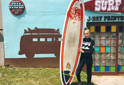 Mädchen mit Surfbrett in der Hand posiert vor einem afrikanischen Surfshop